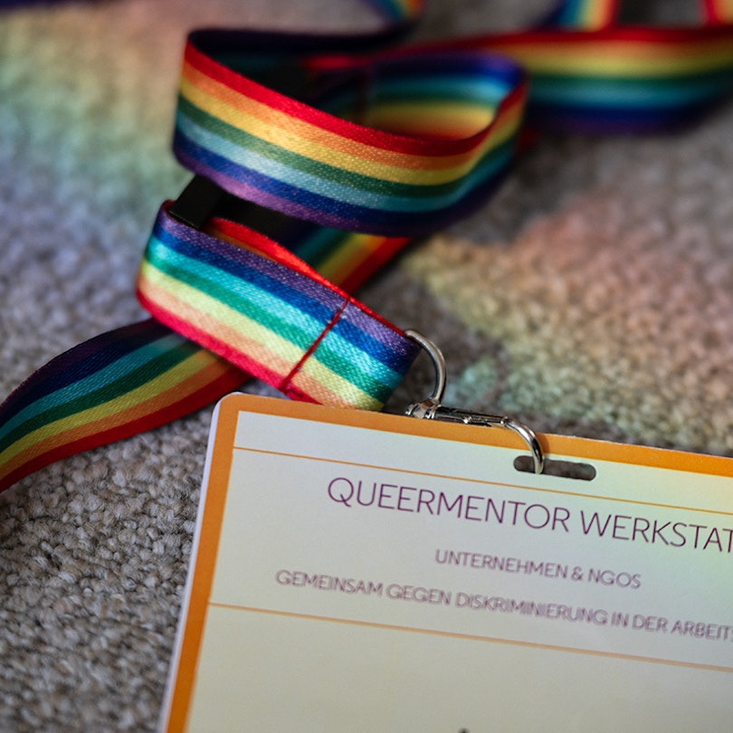 Ein angeschnittenes Badge mit der Aufschrift "Queermentor Werkstatt". Es ist befestigt an einem Regenbogenband