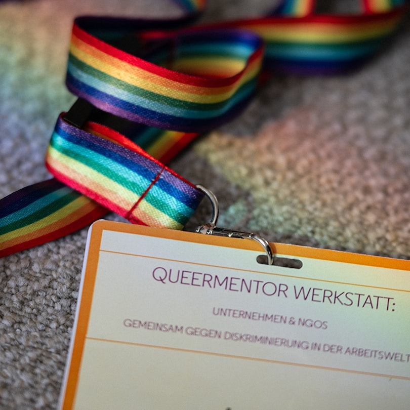 Ein angeschnittenes Badge mit der Aufschrift "Queermentor Werkstatt". Es ist befestigt an einem Regenbogenband