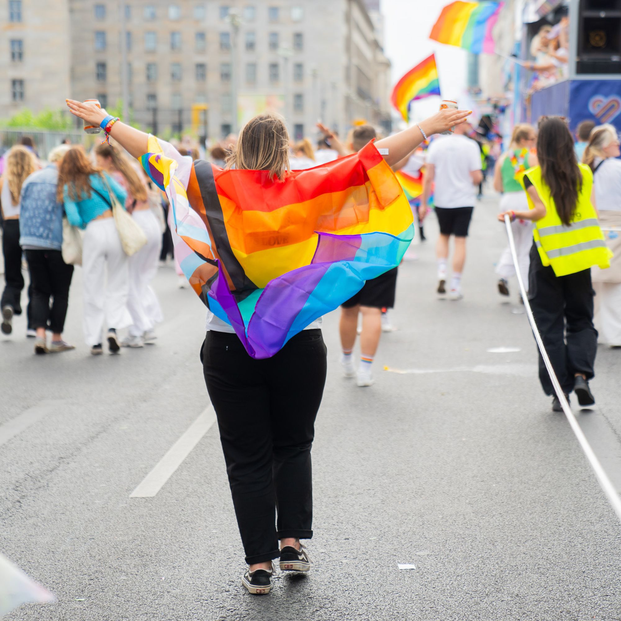 Person mit progressiver Pride-Flagge auf dem Rücken läuft mit ausgebreiteten Armen über eine Straße bei einer CSD-Demonstration. Im Hintergrund sind weitere Teilnehmende und Regenbogenfahnen zu sehen.